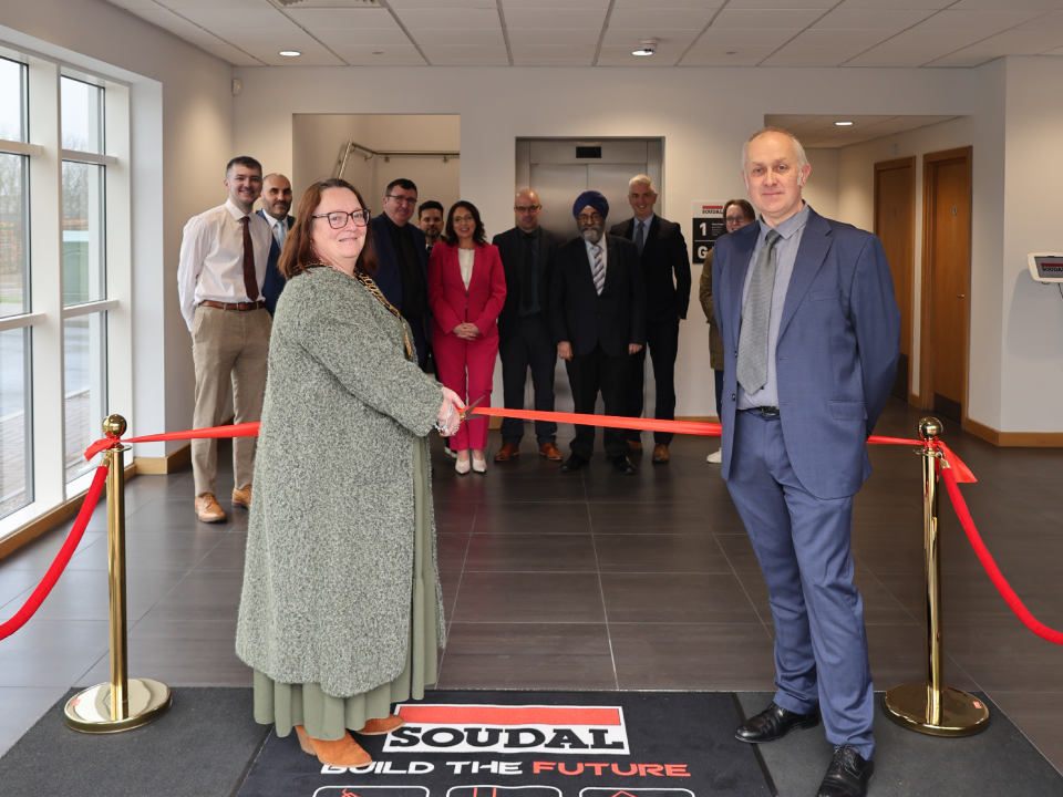 0ac013d5-4fe8-416b-82a5-687da8664889-1 - Soudal India - Soudal India Two people in the foreground cut a red ribbon during a ribbon‑cutting ceremony in a modern lobby, with a group of colleagues watching in the background.