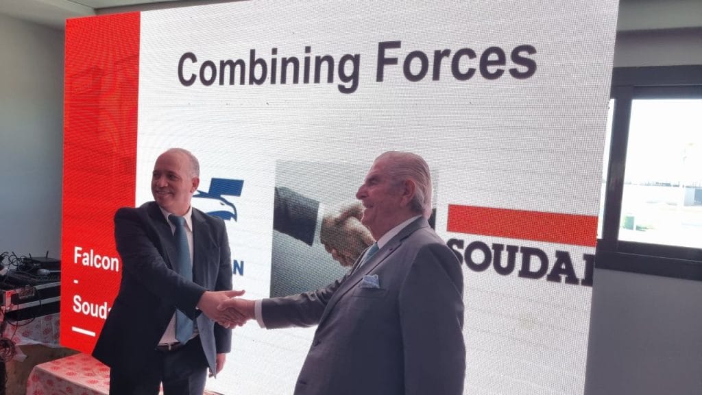 Two men in suits shake hands in front of a large screen that reads 'Combining Forces' with corporate logos in the background.