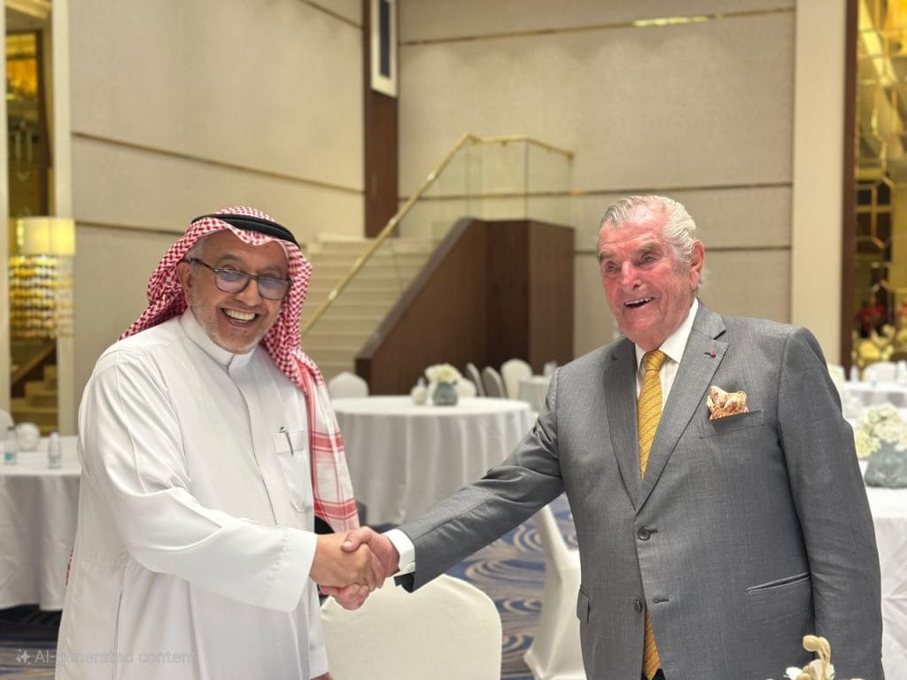 Two men in a handshake at a formal event, one in traditional Arabian attire and the other in a gray suit, smiling in a banquet hall.