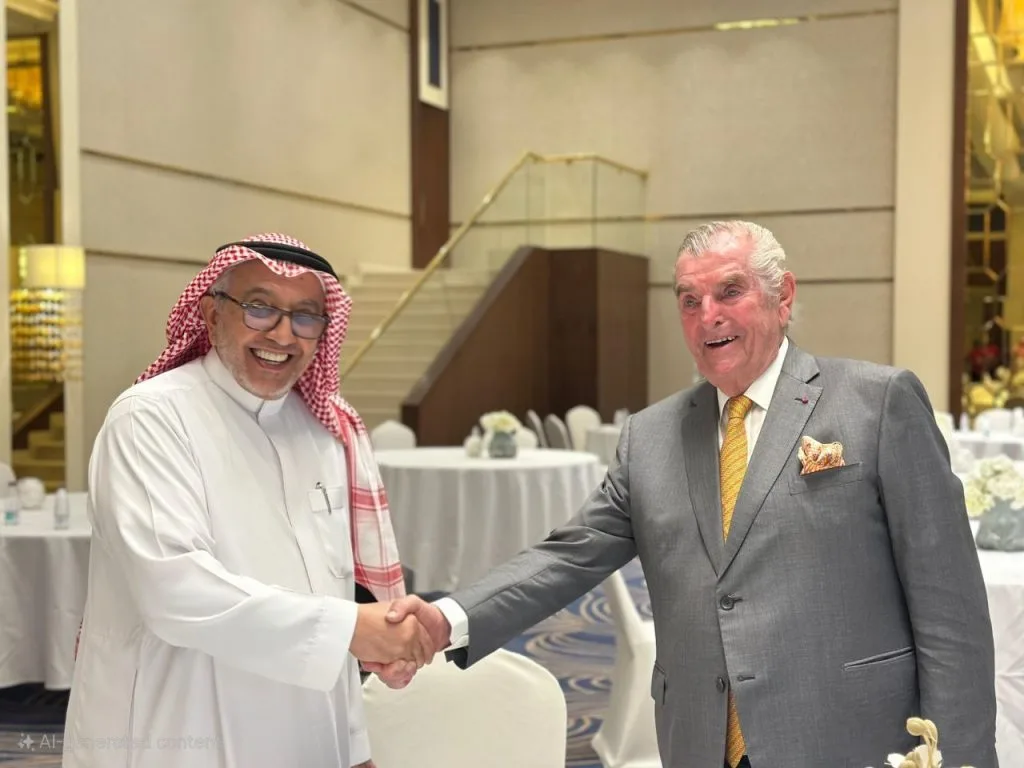 Two men in a handshake at a formal event, one in traditional Arabian attire and the other in a gray suit, smiling in a banquet hall.