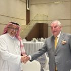 Two men in a handshake at a formal event, one in traditional Arabian attire and the other in a gray suit, smiling in a banquet hall.