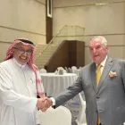 Two men in a handshake at a formal event, one in traditional Arabian attire and the other in a gray suit, smiling in a banquet hall.