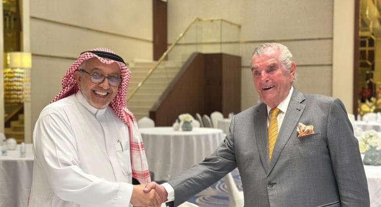 Two men in a handshake at a formal event, one in traditional Arabian attire and the other in a gray suit, smiling in a banquet hall.