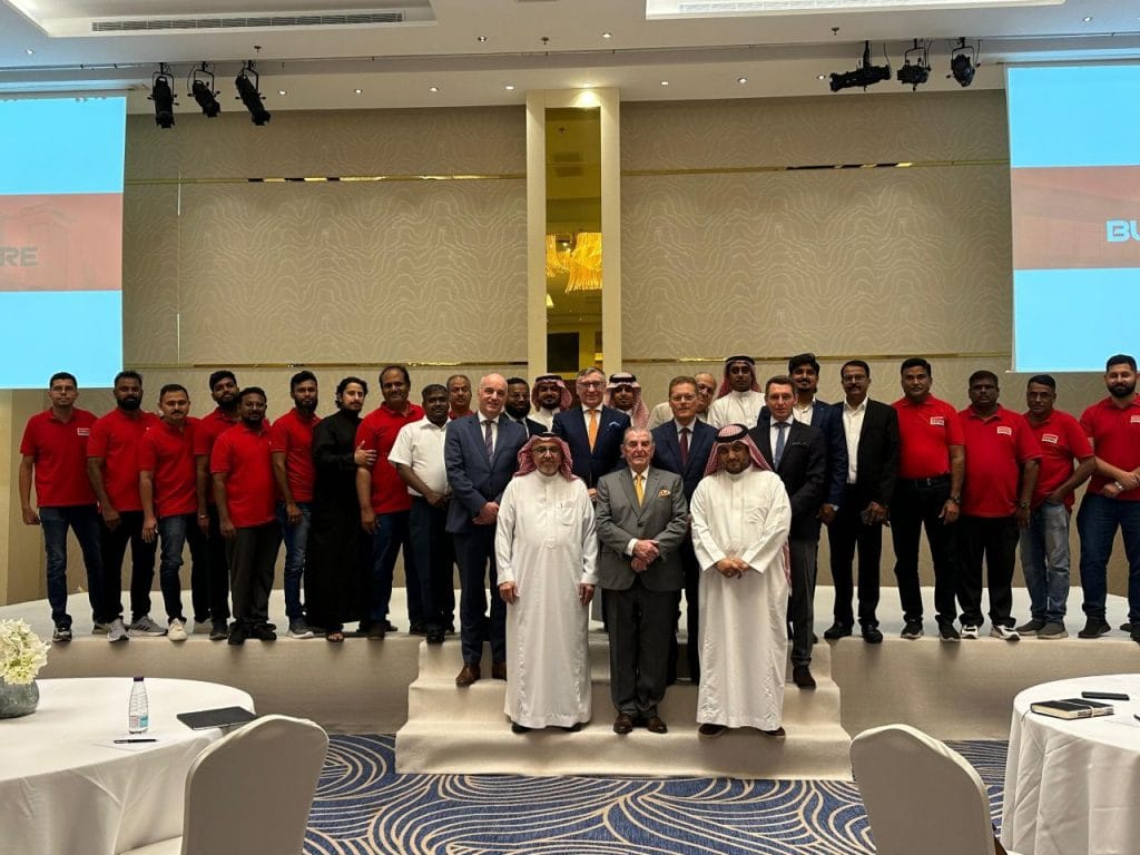 Group of men on a stage posing for a formal photo at a conference hall; many wear red shirts, others in suits or traditional dress.