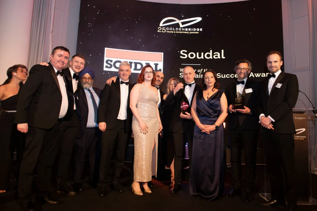 Group of formally dressed people posing on stage at an awards ceremony, with a backdrop reading 'Soudal' and 'Goldenbridge Trade & Investment Awards' behind them.