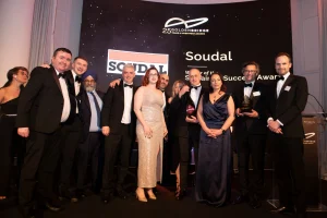 Group of formally dressed people posing on stage at an awards ceremony, with a backdrop reading 'Soudal' and 'Goldenbridge Trade & Investment Awards' behind them.