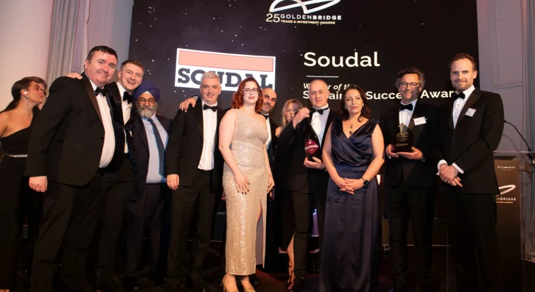Group of formally dressed people posing on stage at an awards ceremony, with a backdrop reading 'Soudal' and 'Goldenbridge Trade & Investment Awards' behind them.