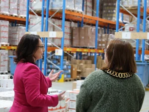 Two women having a discussion in a warehouse with tall blue and orange racks and stacked pallets behind them in the background.