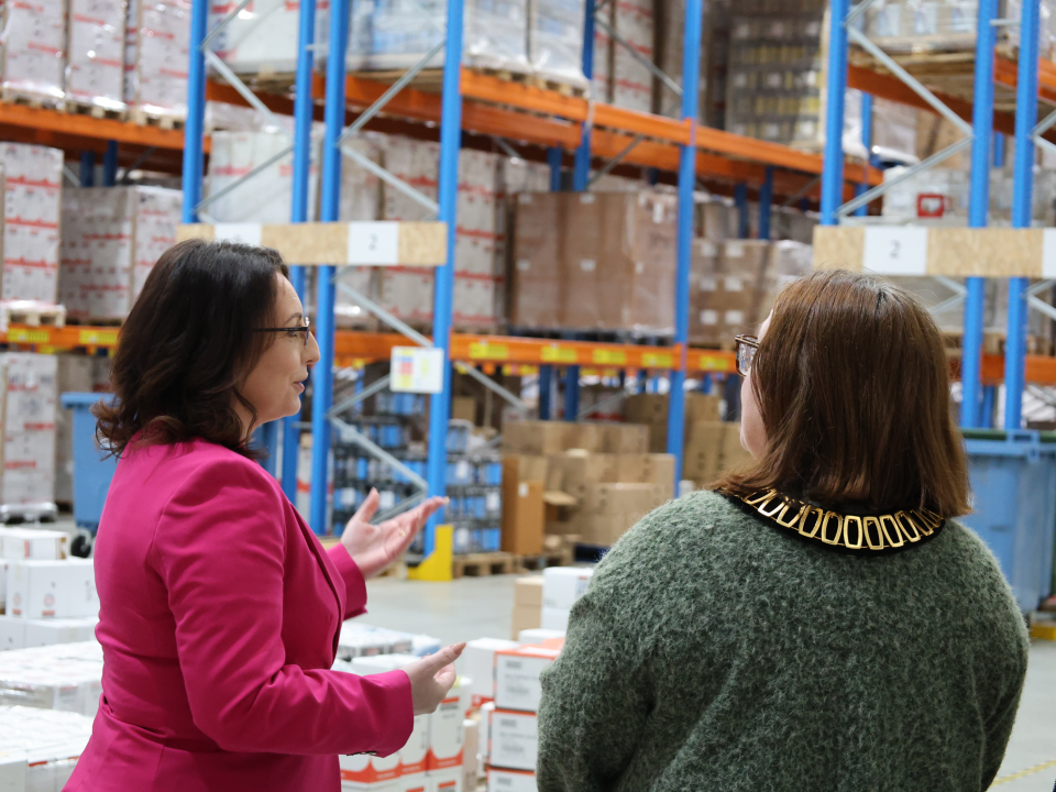 3f56d9dd-581c-4265-8ab1-8f034d80483f - Soudal India - Soudal India Two women having a discussion in a warehouse with tall blue and orange racks and stacked pallets behind them in the background.