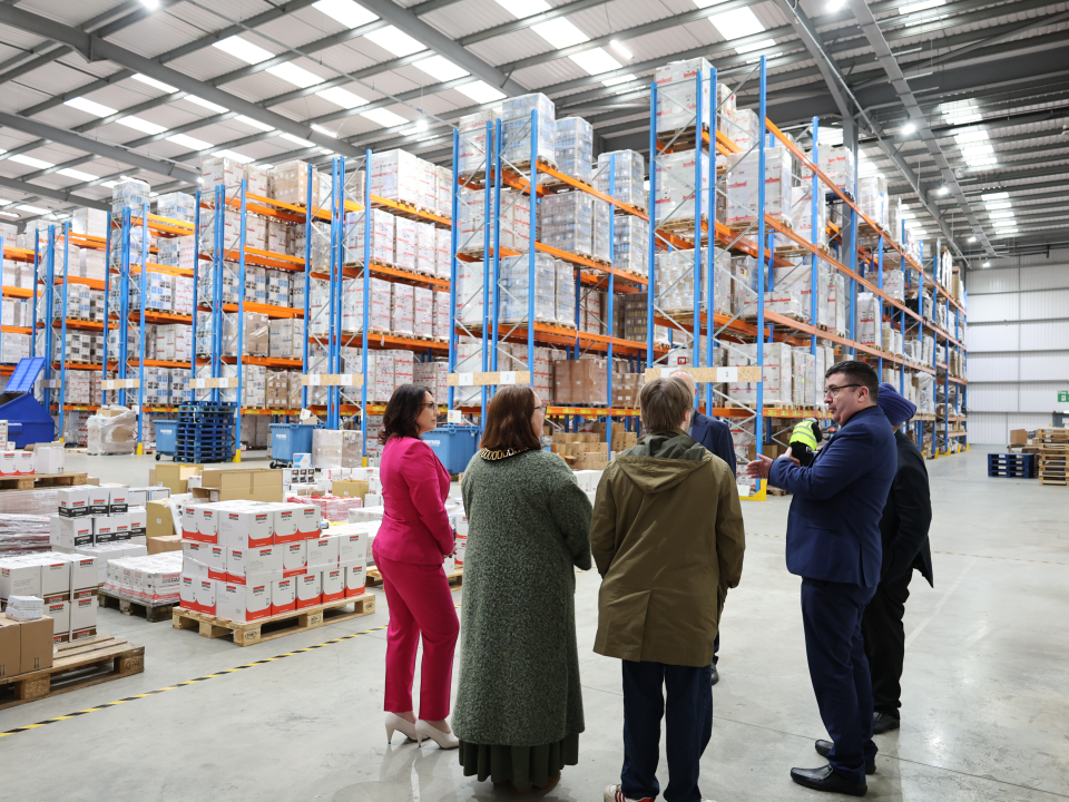 7c783a62-cbe9-49d2-8703-421688fefd1a - Soudal India - Soudal India Group of six coworkers in a warehouse talking in front of tall racks filled with pallets and boxes.